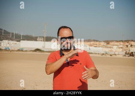 Homme caucasien dans une chemise rouge et des lunettes de soleil à la plage faisant quelques gestes avec ses mains Banque D'Images