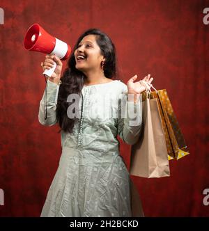 Bonne jeune femme indienne faisant annnouncement sur le mégaphone au sujet des ventes en ligne ou e-commerce en tenant des sacs d'achats - concept de promotion, spécial Banque D'Images