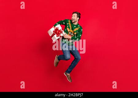 Photo pleine grandeur de sourire heureux bonne humeur gars dans laid pull jump tenir boîte présente isolée sur fond rouge couleur Banque D'Images