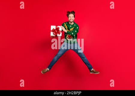 Photo pleine grandeur de gaie homme de bonne humeur sautant célébrer les chrismas tenir boîte présente isolée sur fond rouge vif Banque D'Images