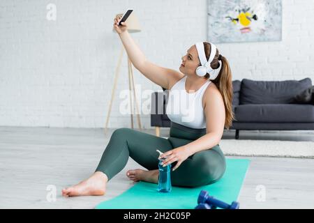 Vue latérale d'une sportswoman souriante et de taille plus grande dans un casque qui prend le selfie sur un smartphone près d'une bouteille de sport sur un tapis de fitness Banque D'Images