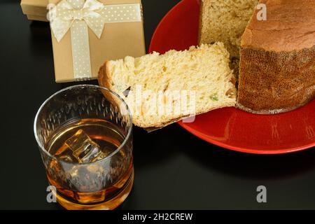 Tranche de panettone sur une assiette rouge, à côté d'un verre de whisky et de glace et d'une boîte cadeau. Banque D'Images