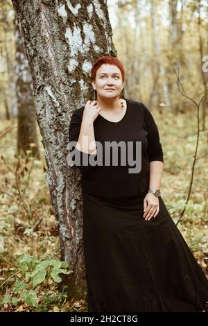 Femme d'âge moyen de 30-40 ans dans la forêt d'automne se dresse par arbre, pensif, rêveur. Banque D'Images