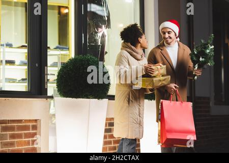 joyeux homme à santa chapeau tenant des sacs de shopping et de pin près de l'afrique de l'amérique petite amie avec des boîtes-cadeaux près du centre commercial Banque D'Images