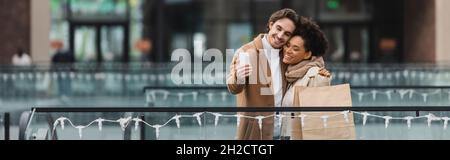 joyeux couple multiethnique avec des tasses en papier et des sacs de shopping prenant selfie dans le centre commercial, bannière Banque D'Images