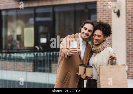 joyeux couple multiethnique avec des tasses en papier et des sacs de shopping emportant le selfie dans le centre commercial Banque D'Images