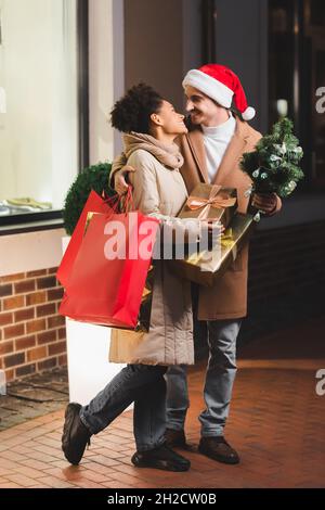 joyeux homme dans le chapeau de santa tenant des sacs d'achats et de sapin près de la petite amie afro-américaine avec des boîtes-cadeaux Banque D'Images