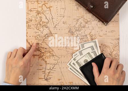 les mains des femmes tiennent un portefeuille en cuir noir avec de l'argent et montrent l'endroit de repos sur la carte du monde. journal marron sur une table en bois blanc Banque D'Images