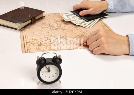 les mains des femmes tiennent un portefeuille en cuir noir avec de l'argent et montrent l'endroit de repos sur la carte du monde. sur une table blanche marron journal et réveil. Banque D'Images