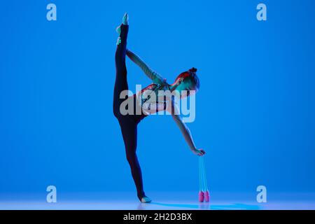 Portrait d'une petite fille, gymnaste rythmique professionnel en train de jouer avec des clubs de gymnastique isolés sur fond bleu au néon Banque D'Images