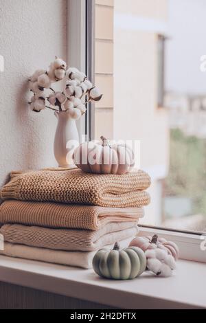 Une pile de chandails tricotés chauds sur le rebord de la fenêtre avec des citrouilles et un vase de coton plante le jour de la pluie, dans des tons pastel. Banque D'Images