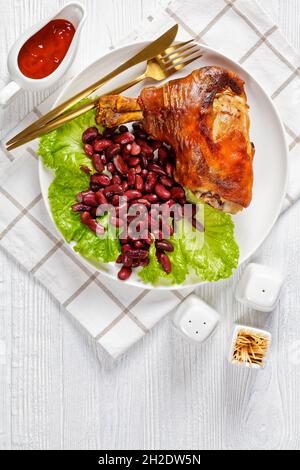tiges de porc grillées croustillantes sur une assiette blanche avec des feuilles de laitue fraîches et des haricots rouges cuits sur une table en bois, vue verticale depuis le dessus Banque D'Images