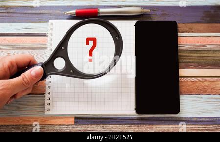 Sur un arrière-plan rayé se trouve un ordinateur portable avec un téléphone.Une loupe avec un point d'interrogation est tenue par la main d'une femme. Banque D'Images