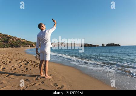 Beau homme heureux en chemise blanche prenant le selfie sur la plage.Concept vacances d'été. Banque D'Images