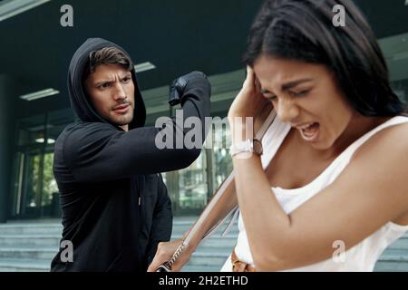 Un voleur agressif veut voler le sac à main d'une fille effrayée. Homme menaçant avec le pistolet et veulent punch jeune femme européenne. Homme bandit porter noir ho Banque D'Images