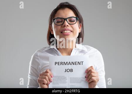 Bonne jeune femme tenant un signe - emploi permanent - avec contrat de travail devant sa poitrine sur un fond gris studio Banque D'Images