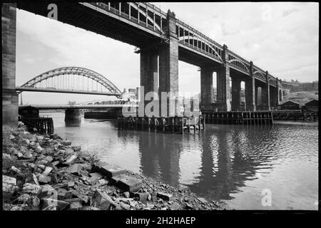 High Level Bridge, Newcastle upon Tyne, c1955-c1980.Vue générale de la rive nord de la rivière Tyne, montrant le pont de haut niveau au premier plan et le pont Swing et le pont de nouveau Tyne en arrière-plan, vus de l'ouest.Le pont de haut niveau est un pont ferroviaire et routier combiné achevé en 1849.La chaussée est sur la poutre inférieure, et les chemins de fer au-dessus, soutenus par six arches segmentaires de fonte, et cinq jetées en pierre.Les deux niveaux ont des parapet ouverts, et le côté sud du pont se compose d'un quai ashlar qui s'étend vers la rivière.La rotation la plus courte se trouve à gauche Banque D'Images