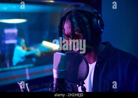 Jeune homme africain en lunettes de soleil avec des dreadlocks portant des écouteurs et chantant pour mettre sa chanson en microphone dans le studio Banque D'Images