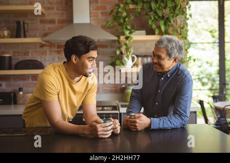 Le fils biracial adulte souriant et le père senior parlent et boivent du café dans la cuisine Banque D'Images