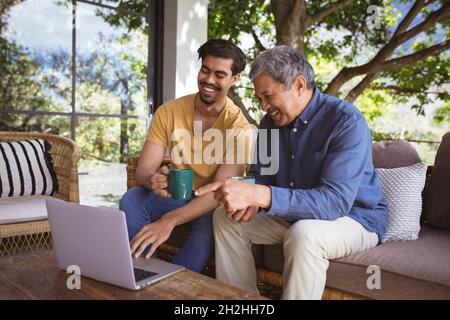 Le fils adulte et le père senior souriant, biracial, utilisent un ordinateur portable et boivent du café dans le jardin Banque D'Images