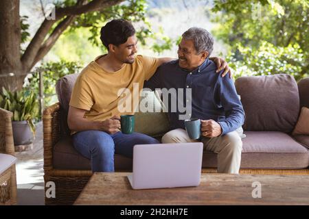 Le fils adulte et le père senior souriant, biracial, s'embrassant et buvant du café dans le jardin Banque D'Images