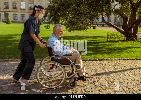 Prise de vue en longueur d'une infirmière soignante avec un homme en fauteuil roulant lors d'une promenade dans le parc près de l'hôpital Banque D'Images