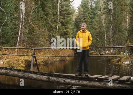 Voyages locaux et randonnées le week-end.Un voyageur d'âge moyen à capuchon jaune se tient sur le pont au-dessus de la rivière dans la forêt, profitant de la belle journée d'automne. Banque D'Images