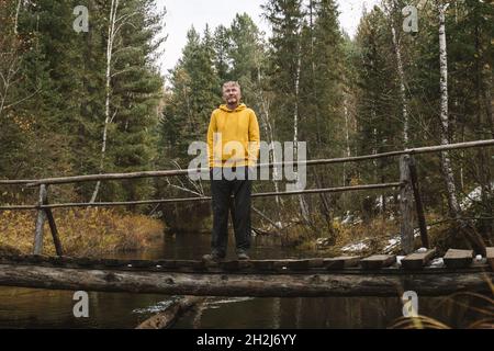 Écotourisme.Voyageur d'âge moyen à capuchon jaune est debout sur le pont au-dessus de la rivière dans la forêt, appréciant belle journée d'automne dehors.Voyages locaux Banque D'Images