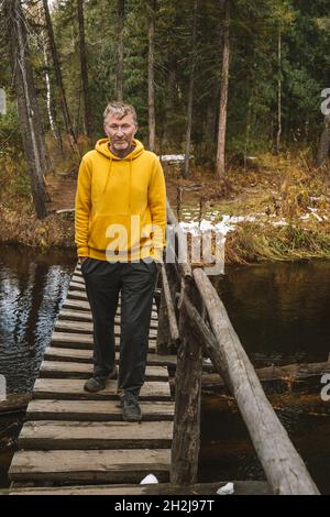 Écotourisme.Un voyageur d'âge moyen à capuchon jaune traverse un pont en bois au-dessus de la rivière dans une forêt d'automne.Randonnées locales et promenades le week-end. Banque D'Images