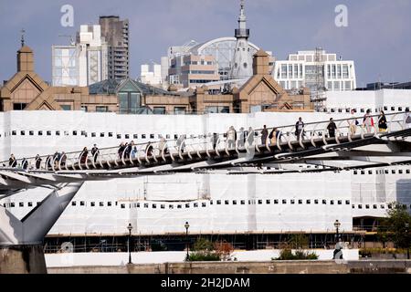 Des bâches dissimulent des travaux de construction sur des immeubles de bureaux au bord de la rivière, tandis que les Londoniens et les visiteurs de la capitale marchent sur le pont du millénaire qui traverse la Tamise, le 19 octobre 2021, à Londres, en Angleterre.La plus récente traversée de la rivière de Londres depuis plus de 100 ans a coïncidé avec le Millenium en 2000.Elle a été achevée à la hâte et ouverte au public le 10 juin 2000, quand environ 100,000 personnes l'ont traversée pour découvrir que la structure oscille tellement qu'elle a été forcée de fermer 2 jours plus tard.Au cours des 18 prochains mois, les concepteurs ont ajouté des amortisseurs pour arrêter son oscillation, mais elle est déjà fragile Banque D'Images