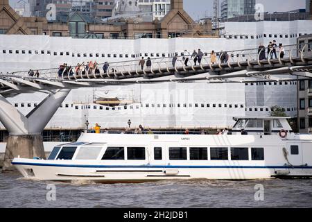 Des bâches dissimulent des travaux sur les bureaux au bord de la rivière, tandis qu'un bateau à pont ouvert passe sous le pont du millénaire qui traverse la Tamise, le 19 octobre 2021, à Londres, en Angleterre.La plus récente traversée de la rivière de Londres depuis plus de 100 ans a coïncidé avec le Millenium en 2000.Elle a été achevée à la hâte et ouverte au public le 10 juin 2000, quand environ 100,000 personnes l'ont traversée pour découvrir que la structure oscille tellement qu'elle a été forcée de fermer 2 jours plus tard.Au cours des 18 prochains mois, les concepteurs ont ajouté des amortisseurs pour arrêter son oscillation, mais elle symbolisait déjà ce qui était embar Banque D'Images