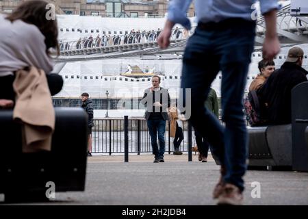 Vu à travers le public marchant le long du fleuve de la Tamise à Bankside, la couverture cache les travaux de construction sur les propriétés de bureau tandis que les Londoniens et les visiteurs de la capitale marchent sur le pont du millénaire qui traverse la Tamise, le 19 octobre 2021, à Londres, en Angleterre.La plus récente traversée de la rivière de Londres depuis plus de 100 ans a coïncidé avec le Millenium en 2000.Elle a été achevée à la hâte et ouverte au public le 10 juin 2000, quand environ 100,000 personnes l'ont traversée pour découvrir que la structure oscille tellement qu'elle a été forcée de fermer 2 jours plus tard.Au cours des 18 prochains mois, les concepteurs ajoutent Banque D'Images