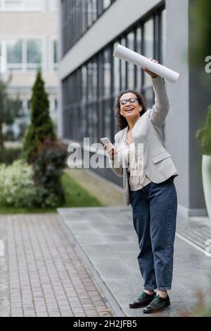 femme d'affaires afro-américaine joyeuse avec téléphone portable qui regarde loin et agite des papiers roulés près d'un bâtiment flou Banque D'Images