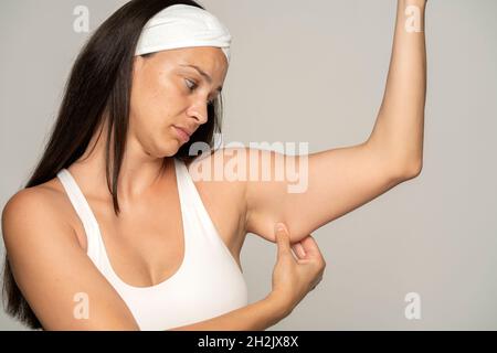 une jeune femme déçue montre la graisse sur son bras sur un fond gris Banque D'Images