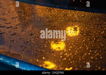 Gros plan d'un raindrop sur le couvercle de coffre d'une voiture la nuit à la lumière des lanternes jaunes Banque D'Images