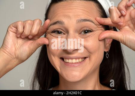 portrait d'une jeune femme souriante pinçant ses rides à côté de ses yeux sur un fond gris Banque D'Images