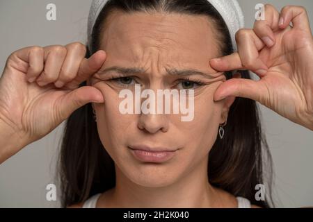 portrait d'une jeune femme triste pinçant ses rides à côté de ses yeux sur un fond gris Banque D'Images