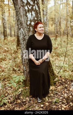 Une femme d'âge moyen de 30-40 ans dans une forêt d'automne se tient près d'un arbre, pensif, rêveur.Photo pleine longueur Banque D'Images