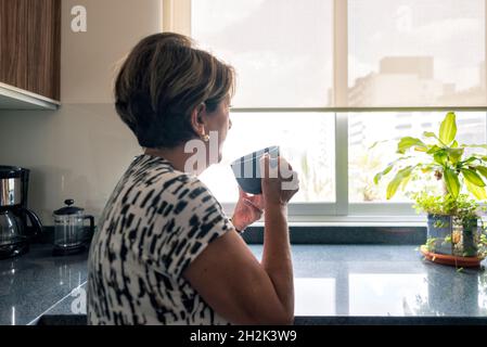 Femme latine mûre regardant la fenêtre boire du café Banque D'Images