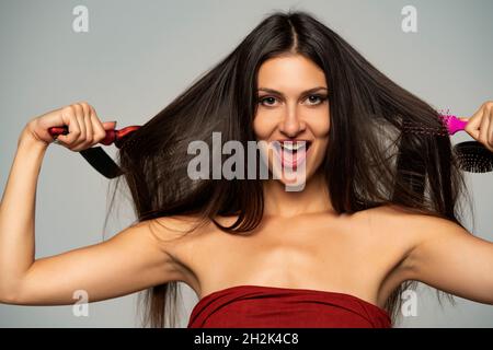 Vue avant de la femme heureuse se brossant ses cheveux avec des brosses à cheveux sur fond gris Banque D'Images
