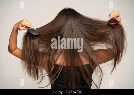 Vue arrière d'une femme se brossant les cheveux avec deux brosses à cheveux sur fond gris Banque D'Images