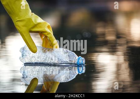 Main avec un gant de protection pour ramasser la bouteille en plastique de l'eau polluée.Nettoyer la nature de la pollution plastique.Questions environnementales Banque D'Images