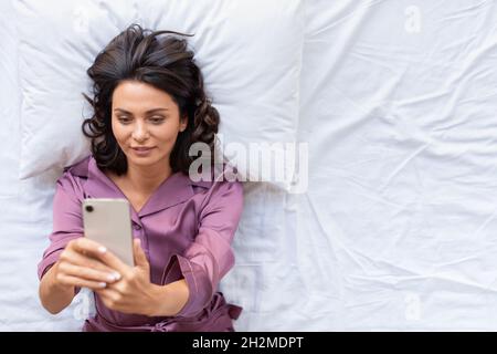 Concept de dépendance aux médias sociaux - vue de dessus d'une femme d'âge moyen qui vérifie les messages et les nouvelles sur les médias sociaux juste après le réveil.Elle se trouve dans le lit et Banque D'Images