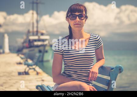 Jeune fille belle avec t-shirt rayé et lunettes de soleil sourire et s'asseoir sur le banc dans la jetée, bateau blanc sur Toroneos bleu golfe turquoise eau dans Halkidiki Banque D'Images