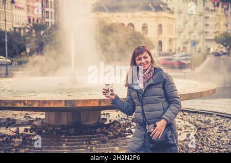 Jeune femme touriste avec le sourire de veste grise et tenir l'eau minérale thérapeutique dans la tasse près de la fontaine source chaude geyser Vridlo dans Karlovy Vary historique Banque D'Images