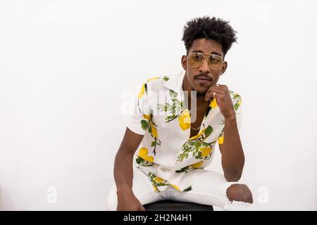 Jeune afro-américain dans une chemise à fleurs et des lunettes de soleil, assis et posant sur un fond blanc Banque D'Images