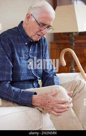 Homme âgé assis sur un canapé à la maison souffrant de douleurs aux genoux causées par l'arthrite Banque D'Images