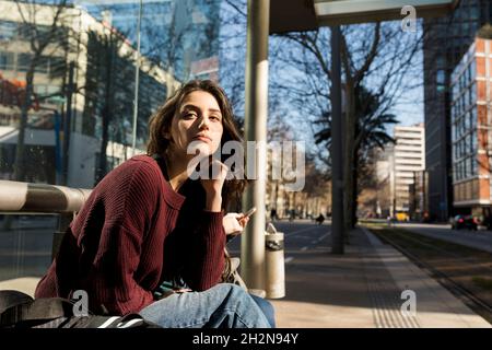 Jeune femme attendant le tram tout en étant assise sur le banc en ville Banque D'Images