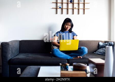 Femme adulte de taille moyenne utilisant un ordinateur portable tout en étant assise à pieds croisés sur un canapé à la maison Banque D'Images