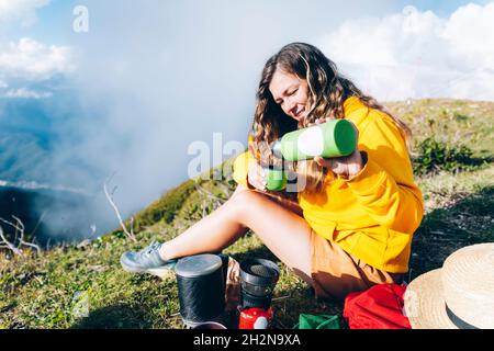 Femme qui verse de la boisson dans un récipient isolé tout en étant assise sur la montagne pendant la journée ensoleillée Banque D'Images